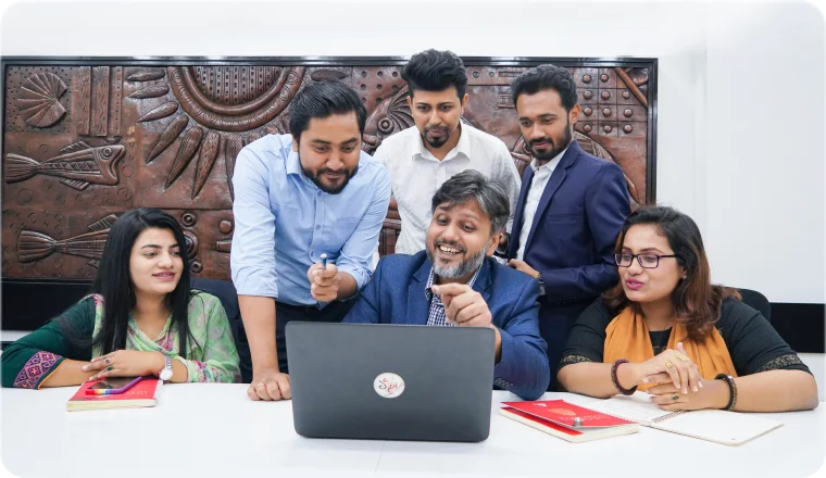 Six members of the Desvert team around a laptop in the studio, smiling as one of them points at the screen.